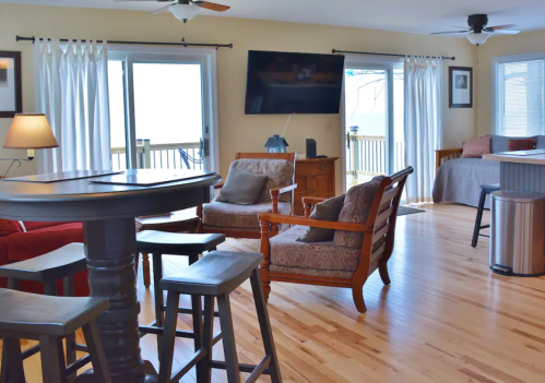Cozy living room with wooden floors, two chairs, a bar table, and large windows overlooking a deck.