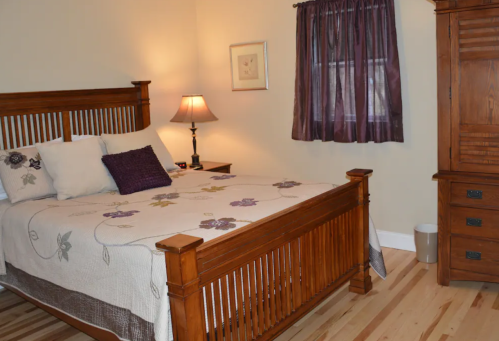 Cozy bedroom with a wooden bed, decorative quilt, lamp, and a dresser against a light-colored wall.