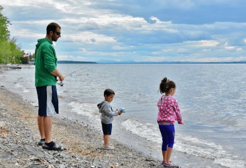 A man and two children fish by a lake, with cloudy skies and a rocky shore in the background.