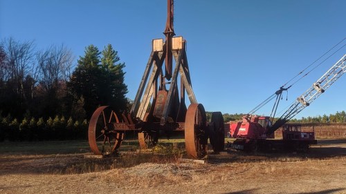 An old, rusted crane with large wheels stands in a field, surrounded by trees under a clear blue sky.