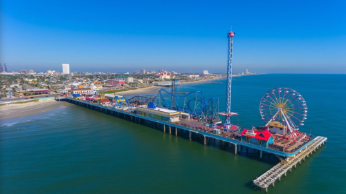 Aerial view of a vibrant amusement pier with rides, games, and ocean in the background under a clear blue sky.