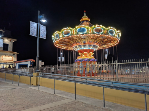 A brightly lit carousel at night, surrounded by a fence, with nearby buildings and streetlights in the background.