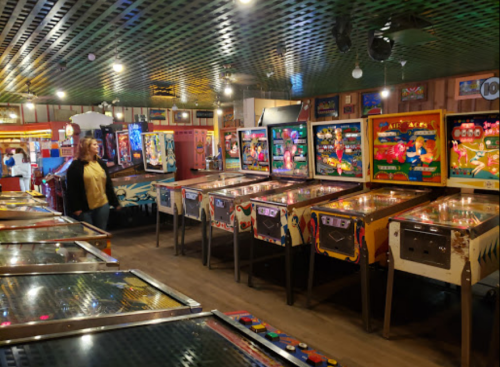 A retro arcade filled with pinball machines and colorful game screens, with a person standing nearby.