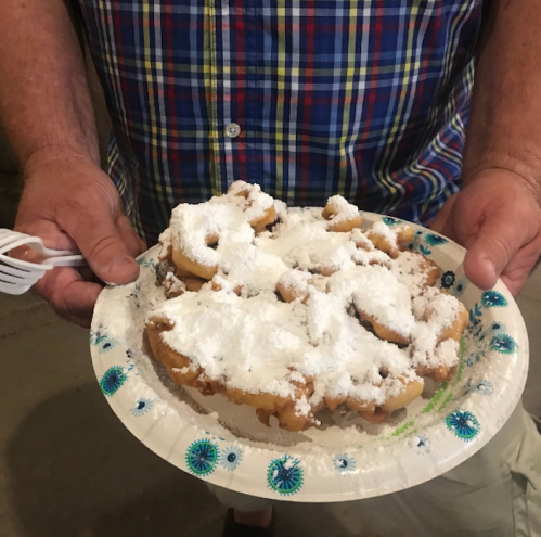 A person holding a plate of funnel cakes dusted with powdered sugar, served on a paper plate.