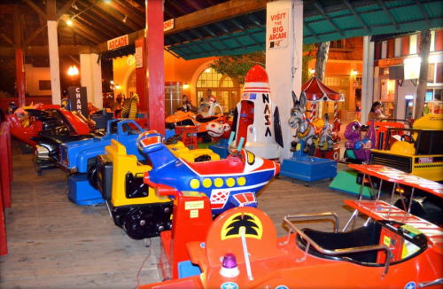 Colorful vintage arcade rides lined up in a lively indoor amusement area. Bright lights and playful decorations visible.