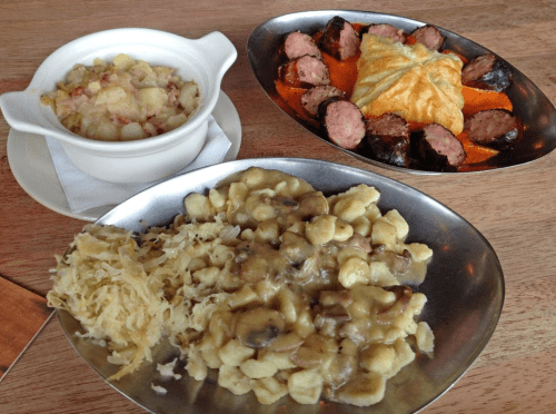 A plate featuring sausage, a pastry, and a side of gnocchi with mushrooms, alongside a bowl of potato salad.