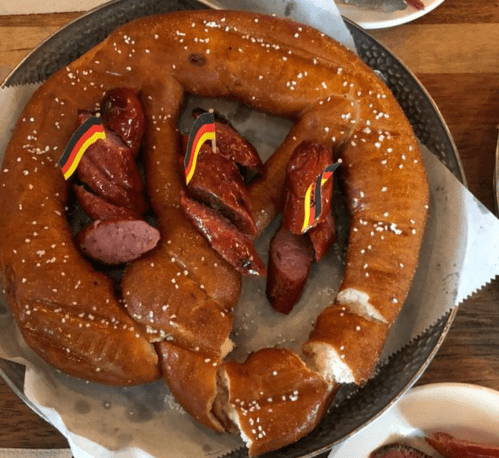A large pretzel with sliced sausages, each topped with small German flags, served on a plate.