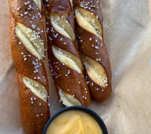 Three twisted pretzel sticks sprinkled with coarse salt, served with a small bowl of cheese dip on a parchment-lined surface.