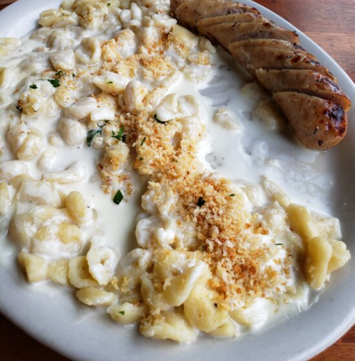A plate of creamy gnocchi topped with breadcrumbs, accompanied by a grilled sausage.