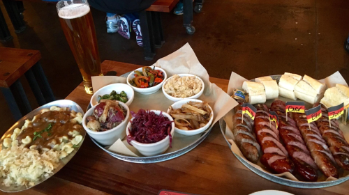 A table spread featuring various sausages, sides like coleslaw and mac and cheese, and a tall glass of beer.