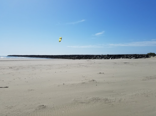 A sandy beach with a clear blue sky and a kite surfer in the distance near a rocky jetty.