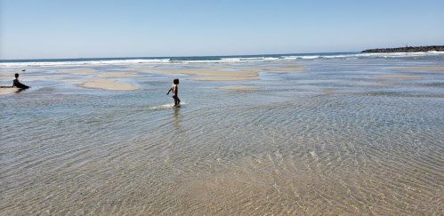 A child walks through shallow water on a beach, with gentle waves and a clear blue sky in the background.