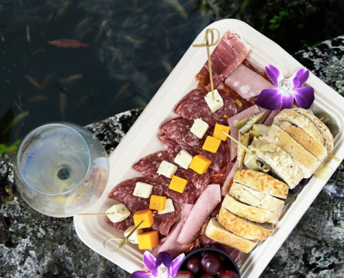 A platter of assorted meats and cheeses garnished with flowers, alongside a glass of drink, set near water.