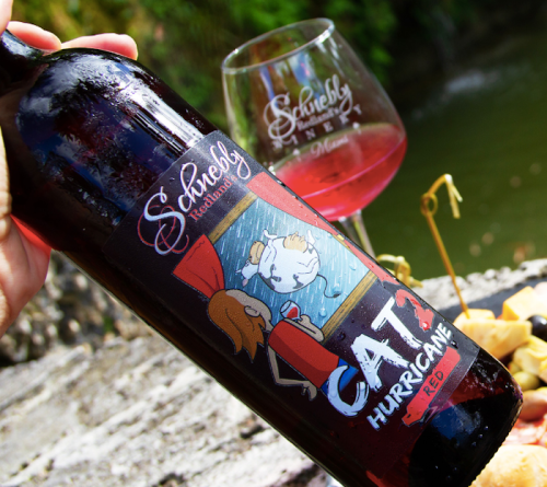 A hand holds a bottle of "Cat Hurricane Red" wine next to a glass, with a scenic background and a spread of food.