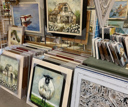 A display of various framed artworks, including landscapes and a sheep, in a gallery or shop setting.