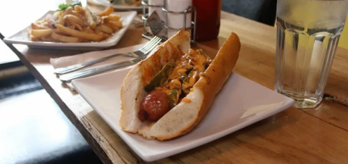 A hot dog in a bun topped with sauce and jalapeños, served on a white plate with a glass of water in the background.