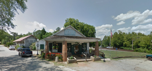A quaint café with a porch, surrounded by greenery and flowers, located on a quiet street with a few parked cars.