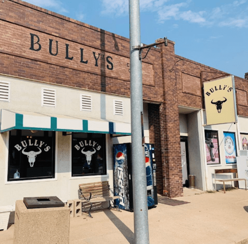 Exterior of Bully's restaurant, featuring a brick facade, large windows, and a striped awning. Clear blue sky above.