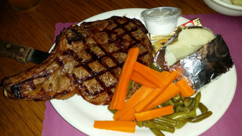 A grilled steak on a plate with carrots, green beans, a baked potato, and a small cup of sauce.