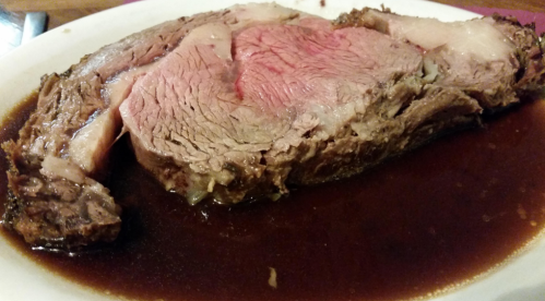 A slice of medium-rare prime rib resting in a rich, dark gravy on a white plate.