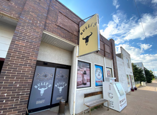Exterior of Bully's bar with a sign, large windows, and an ice vending machine on the sidewalk. Blue sky above.