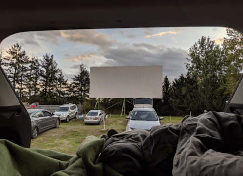 View from a car trunk at a drive-in movie theater, featuring a blank screen and parked cars under a cloudy sky.