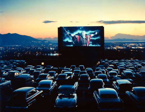 A vintage drive-in theater at sunset, with classic cars parked and a large screen displaying a dramatic scene.