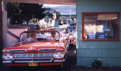 A vintage red convertible car at a drive-in, with two people inside and a retro building in the background.