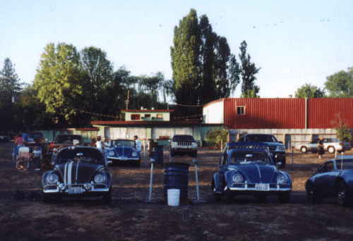 A parking lot with several vintage cars, including two blue Beetles, and a building in the background under a clear sky.