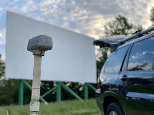 A car parked at a drive-in theater with a blank movie screen in the background under a cloudy sky.