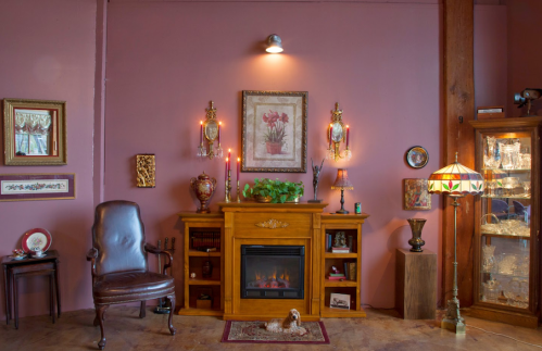 Cozy room with a wooden fireplace, vintage decor, a chair, and a glass display cabinet against a pink wall.