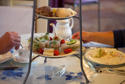 A tiered stand with sandwiches, scones, and desserts, with two hands reaching for the food at a table setting.