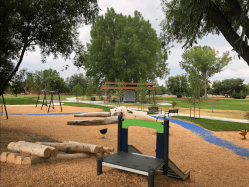 A playground with swings, logs, and a climbing structure, surrounded by trees and a walking path.
