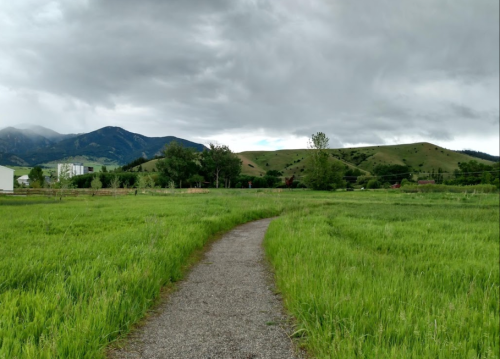 A gravel path winds through lush green fields, with mountains and cloudy skies in the background.