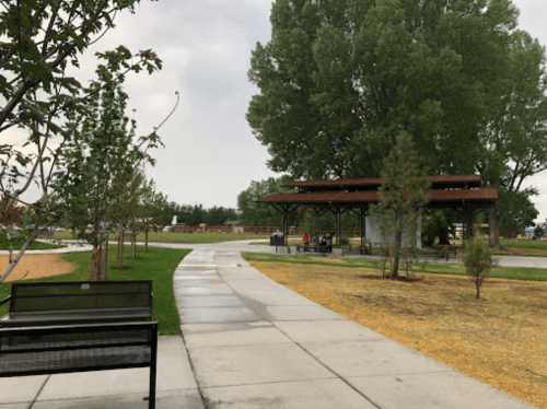 A park pathway lined with trees, leading to a covered pavilion and grassy areas under a cloudy sky.