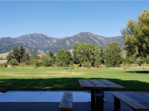 A scenic view of mountains in the background, with a grassy field and a wooden picnic table in the foreground.