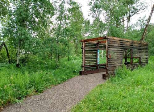 A wooden structure with slatted walls sits beside a gravel path, surrounded by lush greenery and trees.