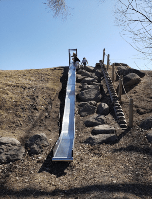Children climbing up a grassy hill to a metal slide, with rocks and a wooden ladder nearby under a clear blue sky.