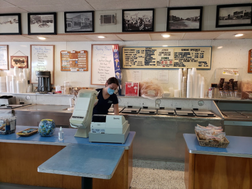 A worker in a retro ice cream shop prepares orders at the counter, surrounded by vintage decor and menu boards.