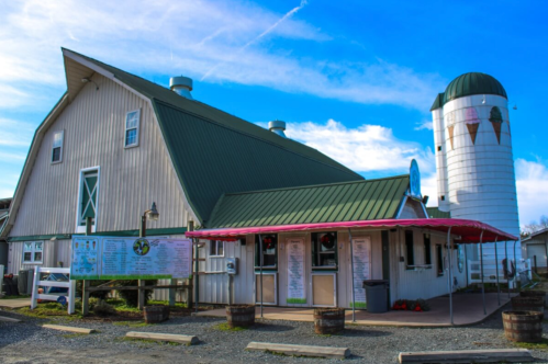 A rustic barn with a green roof and a silo, featuring a menu board outside and a clear blue sky above.
