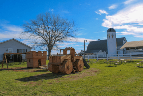 A wooden tractor playground structure beside a barn and a silo under a blue sky with scattered clouds.