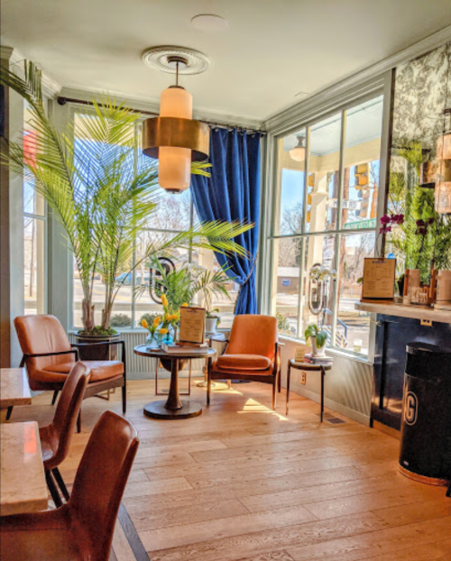Bright café interior with palm plants, two brown chairs, a round table, and large windows letting in natural light.