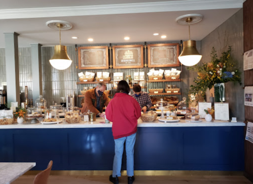 A cozy café interior with a counter displaying baked goods and two staff members assisting a customer.