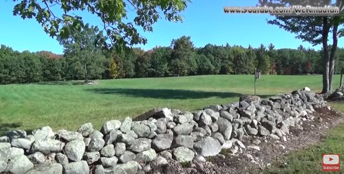 A scenic view of a grassy field bordered by a stone wall, surrounded by trees in autumn colors under a clear blue sky.