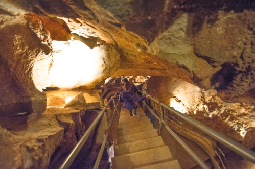 A person descends a staircase in a dimly lit cave, surrounded by rocky walls and illuminated by soft lights.