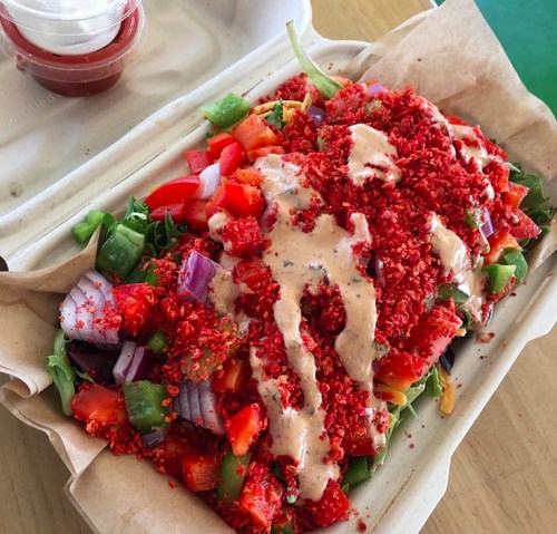 A colorful salad topped with crushed red snacks and drizzled with a creamy dressing, served in a takeout container.
