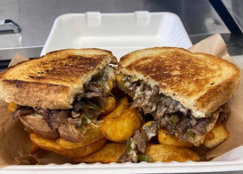 A toasted sandwich filled with beef, peppers, and melted cheese, served with crispy potato wedges on the side.