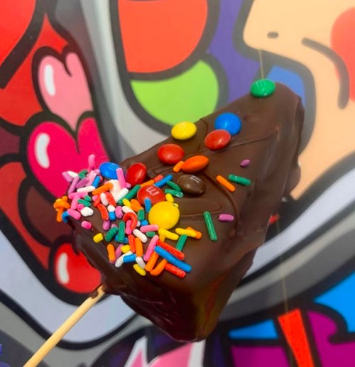 A chocolate-covered triangular treat topped with colorful sprinkles and candies, set against a vibrant background.