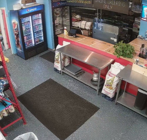 Interior of a pizza shop featuring a counter, shelves, a soda fridge, and bags of ingredients on the floor.
