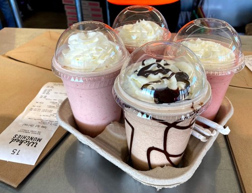 Four milkshake cups with whipped cream, two pink and one brown with chocolate drizzle, in a cardboard tray.